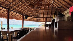 Un área de comedor al aire libre con techo de paja, mesas y sillas de madera, con vista a una playa de aguas turquesas cristalinas.