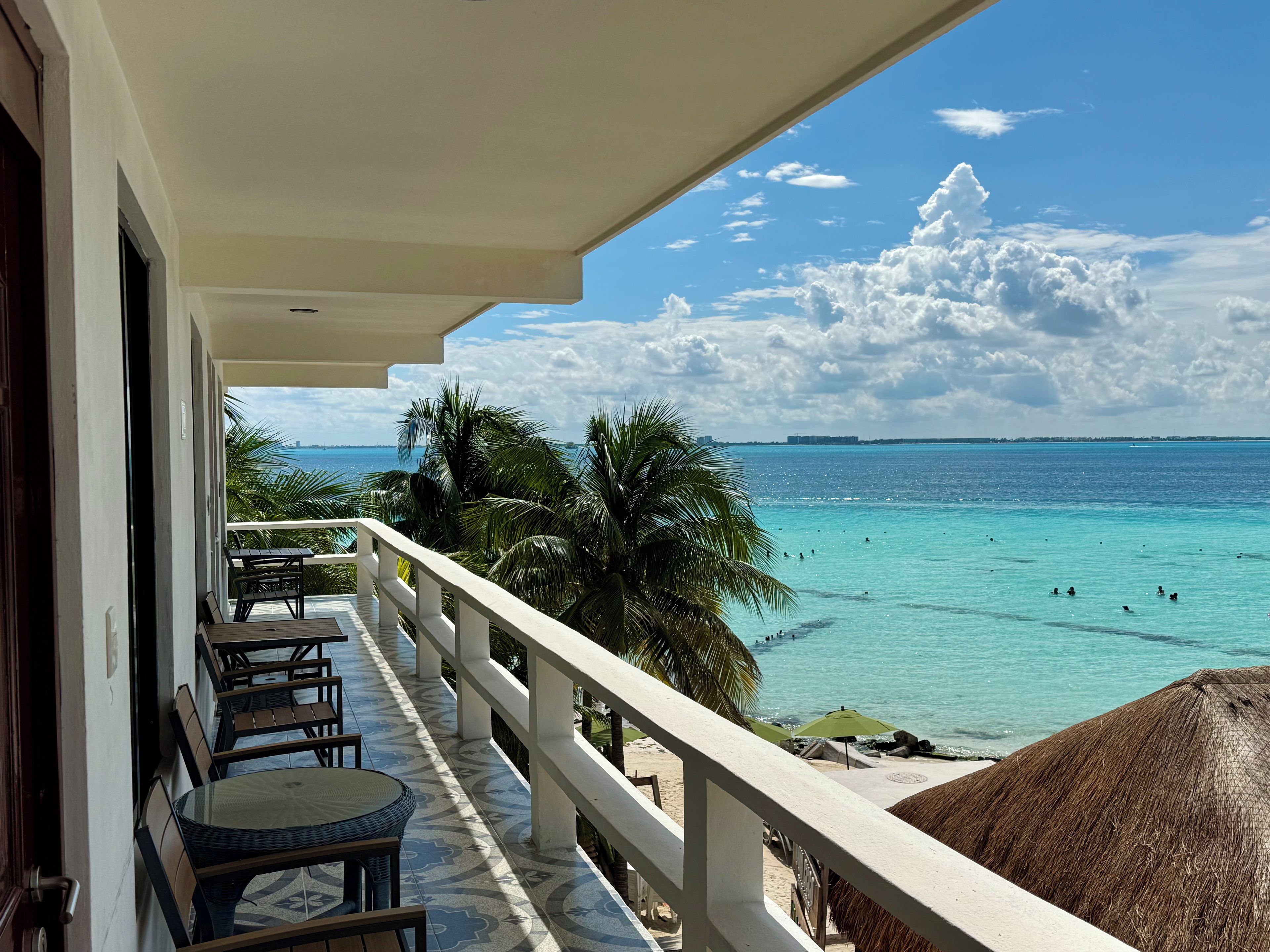 A balcony with a view of the ocean, palm trees, and a thatched umbrella.