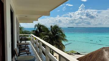 A balcony with a view of the ocean, palm trees, and a thatched umbrella.