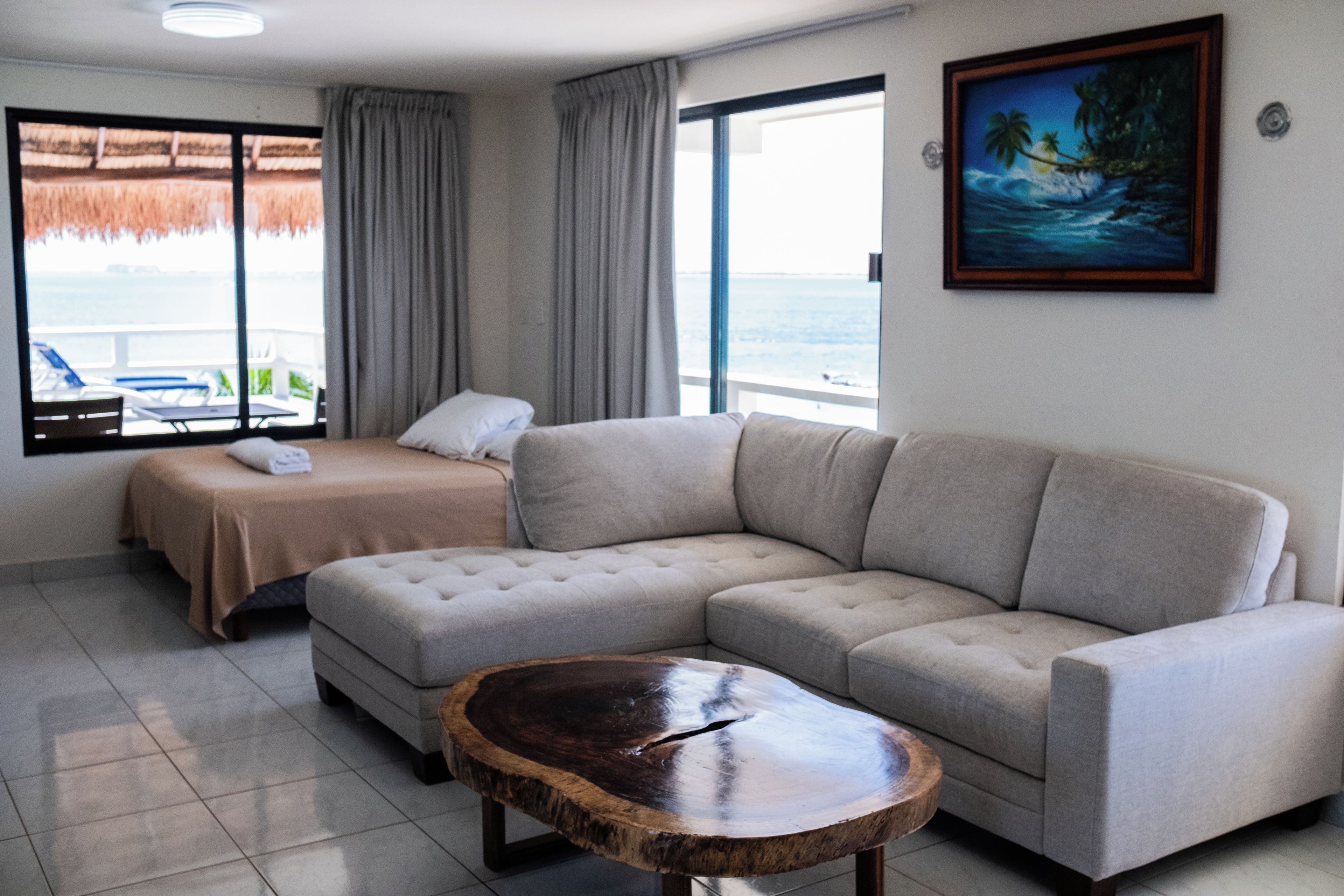 A modern hotel room with a large bed, a sofa, a wooden coffee table, and a painting of a tropical beach.