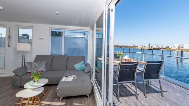 A modern living room with a grey sofa, a small round table, and a balcony with a table and chairs overlooking a body of water. - Awesome ship in Olpenitz with sauna (Olpenitz)