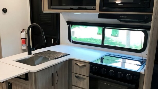 Une cuisine compacte pour camping-car, équipée d’un évier, d’une cuisinière, d’un four à micro-ondes et d’une fenêtre offrant une vue sur la verdure environnante.