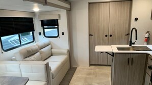 Un intérieur de camping-car compact comprenant un canapé en cuir blanc, une petite table à manger, un coin cuisine avec un évier et deux fenêtres équipées de stores.