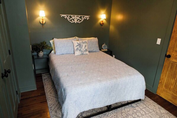 Una cama bien tendida con una manta gris y almohadas, dos lámparas fijadas a la pared, un adorno en la pared, una planta pequeña en una mesita de noche y una puerta de madera.
