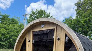 Ein hölzernes, fassförmiges Gebäude mit großer offener Tür und Fenstern, gelegen auf einer Wiese mit Bäumen im Hintergrund.