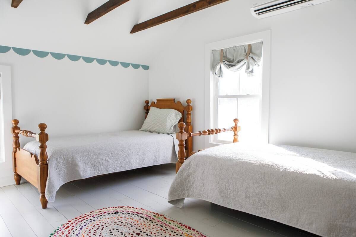 A bedroom with two wooden beds, a white floor, a window with curtains, and a decorative wall hanging.