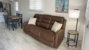 Een woonkamer met een bruine bank, een eettafel met stoelen, een lamp en een schilderij aan de muur.
