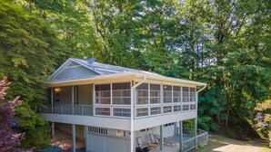 A two-story cabin with a covered porch and a deck, surrounded by trees.