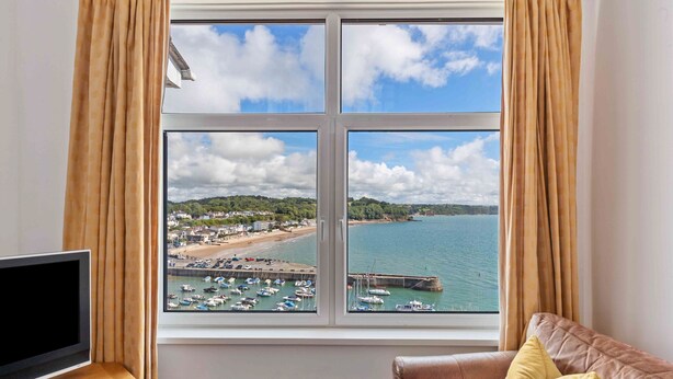 A room with a large window offering a view of a harbor, boats, and a beach. - Captains View - Sea and Harbour Views Parking (Saundersfoot)
