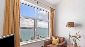 A room with a large window, a television, a sofa, a small table, and a lamp. - Captains View - Sea and Harbour Views Parking (Saundersfoot)