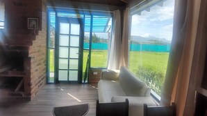 Een knus vertrek met een houten plafond, een bank, een tafel en uitzicht op een groen landschap door grote ramen.