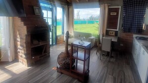 Een knus houten interieur met een stenen openhaard, houten balken in het plafond, een eethoek en uitzicht op een groen landschap door grote ramen.