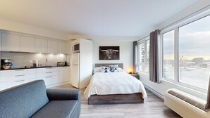Une chambre moderne avec un grand lit, un canapé gris, un coin cuisine et une vue sur la ville grâce à de grandes fenêtres.