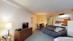 A compact hotel room with a sofa, a TV, a dining table with chairs, a kitchen area with a microwave, and a patterned floor.