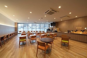 Amplo espaço interno com várias mesas e cadeiras, um balcão com vitrine e grandes janelas que permitem entrada de luz natural.
