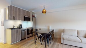 Et kompakt køkken med moderne hårde hvidevarer, et spiseområde med bord og stole, samt en sofa i en opholdsstue.