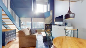 Un apartamento moderno con una escalera, un área de estar con sofá y mesa de centro, una mesa de comedor y una cocina con lámpara colgante.