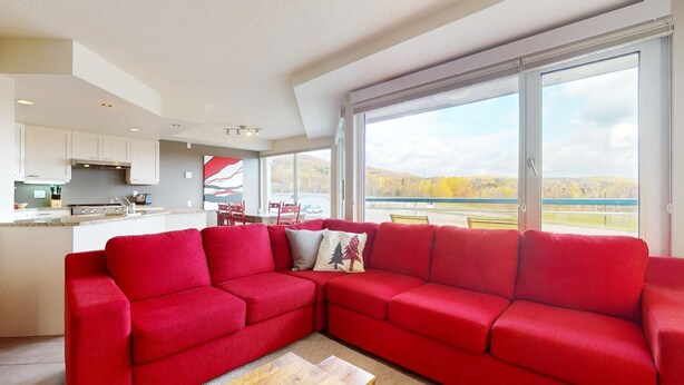 A modern living room with a red sectional sofa, a dining area with a red table and chairs, and large windows offering a view of a lake and mountains.