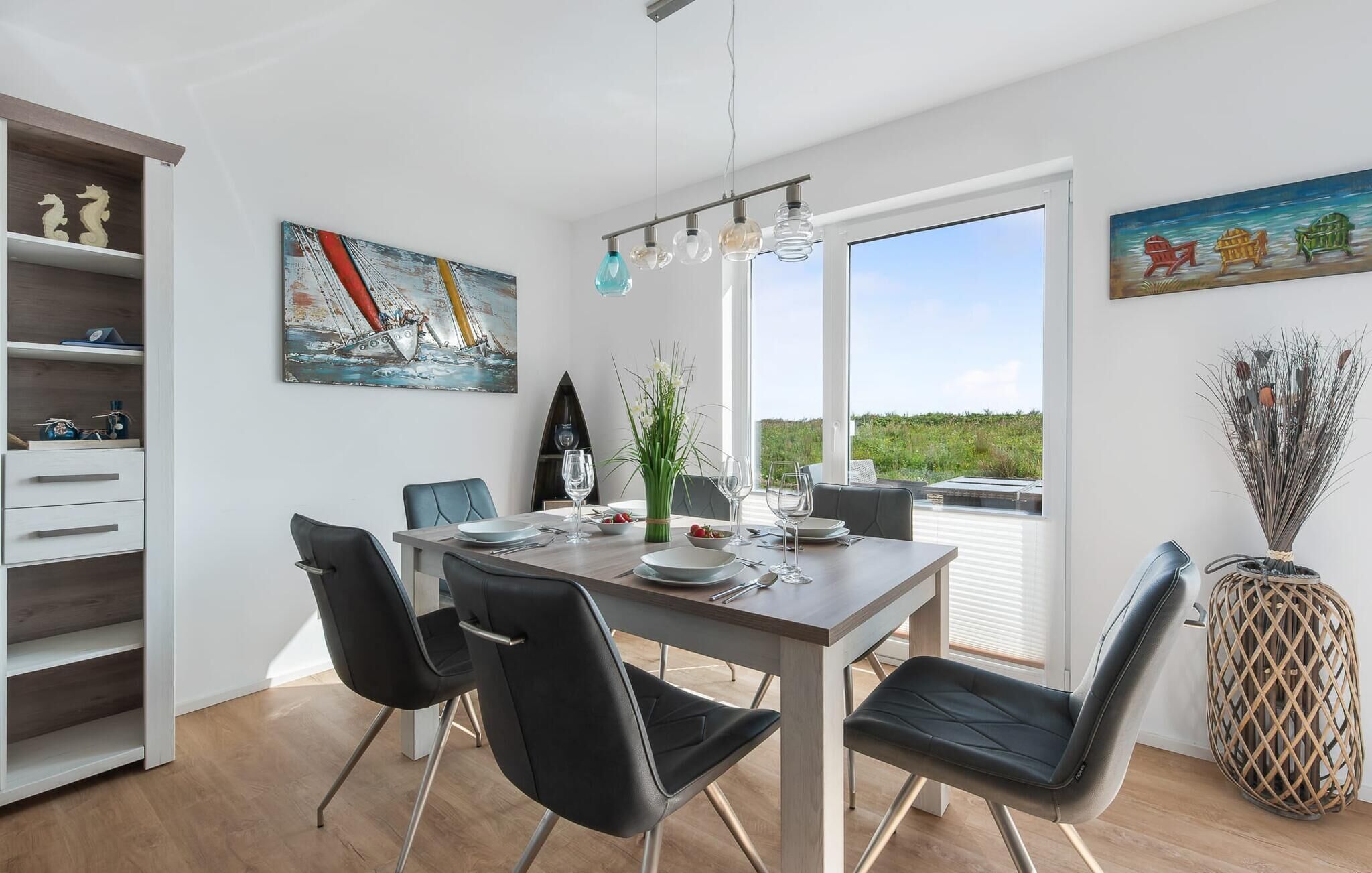 Un coin repas avec une table dressée pour quatre personnes, des chaises, un tableau représentant un voilier et une vue par la fenêtre.
