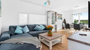 En moderne stue med en grå sofa, et sofabord med en skål frugt, og et spiseområde med et bord og stole.