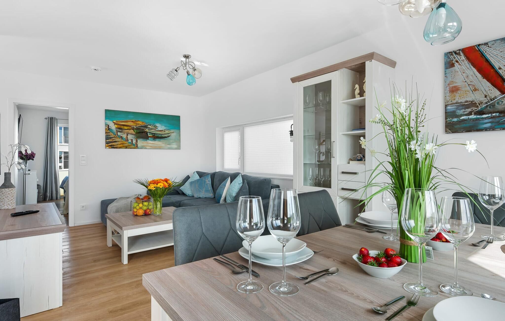 Una sala de estar moderna con un sofá gris, una mesa de comedor dispuesta con copas de vino y platos, y un cuadro con barcos en la pared.