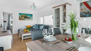 En moderne stue med en grå sofa, et spisebord dækket med vinglas og tallerkener, og et maleri af både på væggen.