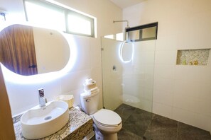 A modern bathroom with a large oval mirror, a freestanding sink, a toilet, and a glass-enclosed shower.