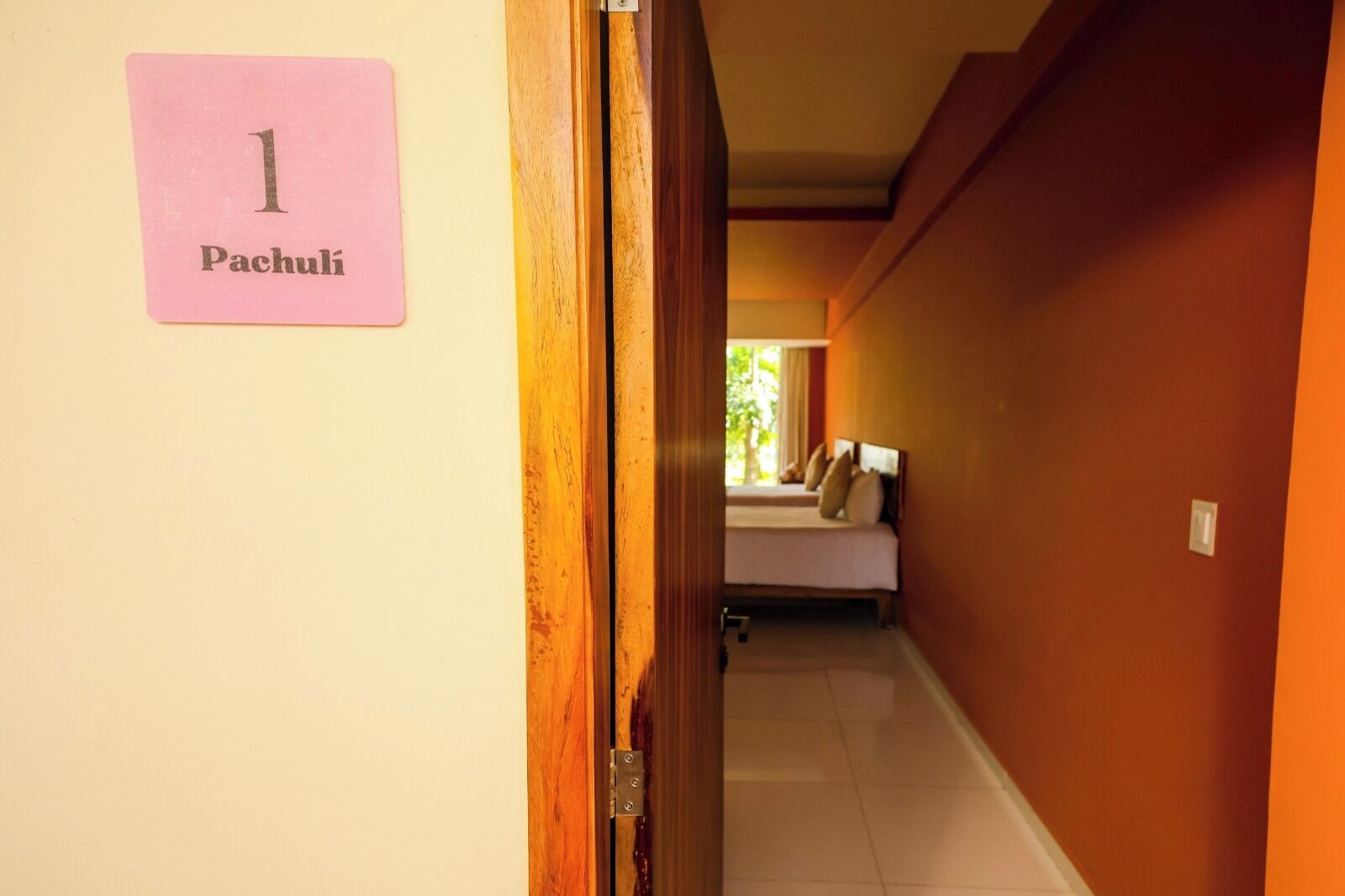 A hallway with a sign reading "1 Pachuli" on the left, leading to a room with a bed and a window.