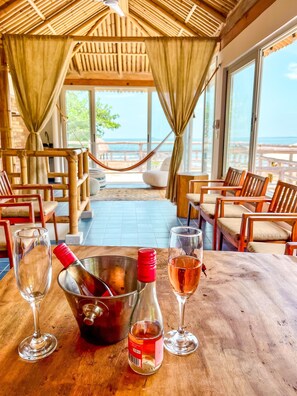 Ein Holztisch mit einer Weinflasche, einem Weinglas und einem Eimer mit Flaschenöffner. Der Tisch steht in einem Raum mit Holzsesseln und Blick auf das Meer durch große Fenster.