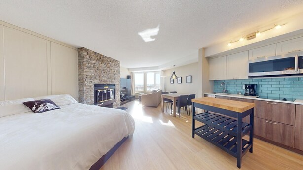 A modern hotel room with a large bed, a stone fireplace, a dining area, and a kitchenette.