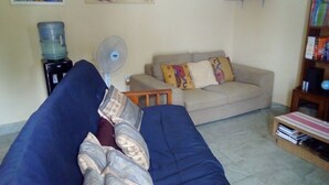 Una sala de estar con un sofá azul, un sofá beige, un dispensador de agua, un ventilador y una pequeña mesa de madera con libros.