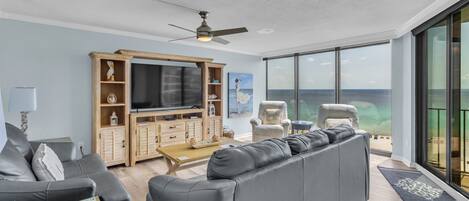 Una sala de estar moderna con un televisor de pantalla plana grande, un mueble de madera para equipo multimedia, un sofá gris y vista al océano a través de amplias ventanas.