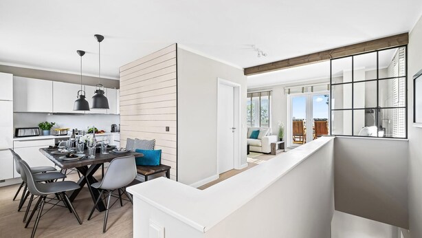 Une cuisine moderne avec des meubles blancs, un coin repas avec une table et des chaises en bois, et une vue sur le salon grâce à une porte ouverte.