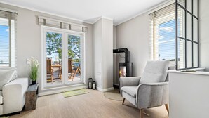 Un salon moderne avec un fauteuil gris, un canapé blanc et une table basse en bois. Une porte-fenêtre coulissante donne sur une terrasse extérieure équipée d’une table et de chaises. On aperçoit une cheminée noire dans un coin.