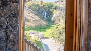 Un muro de piedra y una ventana con marco de madera que da a un jardín, una terraza de madera y una cascada.
