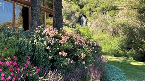 Una cabaña rústica con paredes de piedra y amplias ventanas, rodeada de exuberantes jardines con diversas plantas en flor.