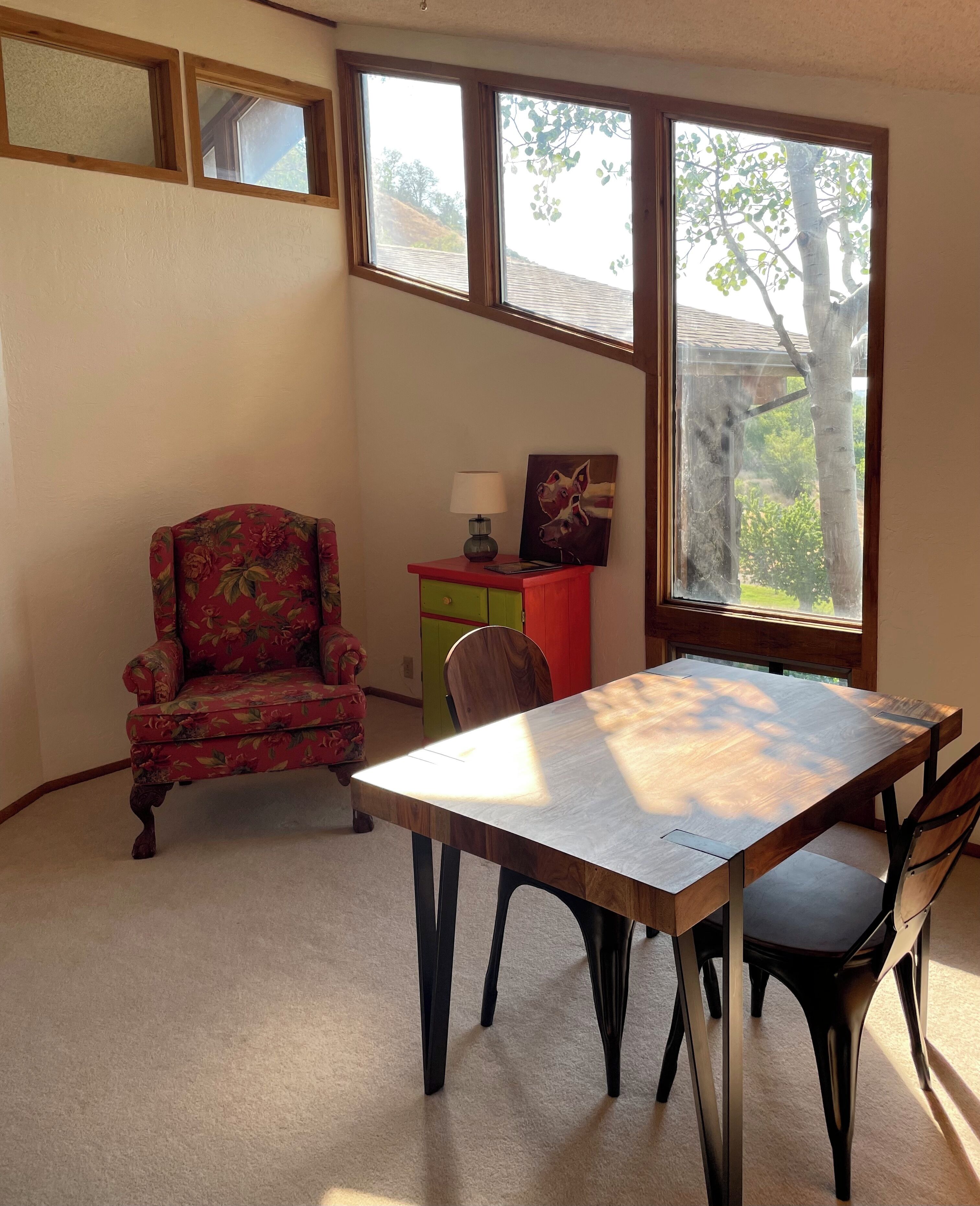 A dining area with a wooden table and chairs, a floral armchair, a small red cabinet with a lamp, and a painting of a dog.