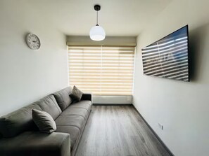 Ein modernes Wohnzimmer mit einer Couch, einer Wanduhr, einer Deckenleuchte und einem großen Fenster mit Jalousien.