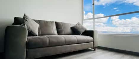 Un salon moderne avec un canapé gris, une horloge au mur et une grande fenêtre donnant sur le ciel.