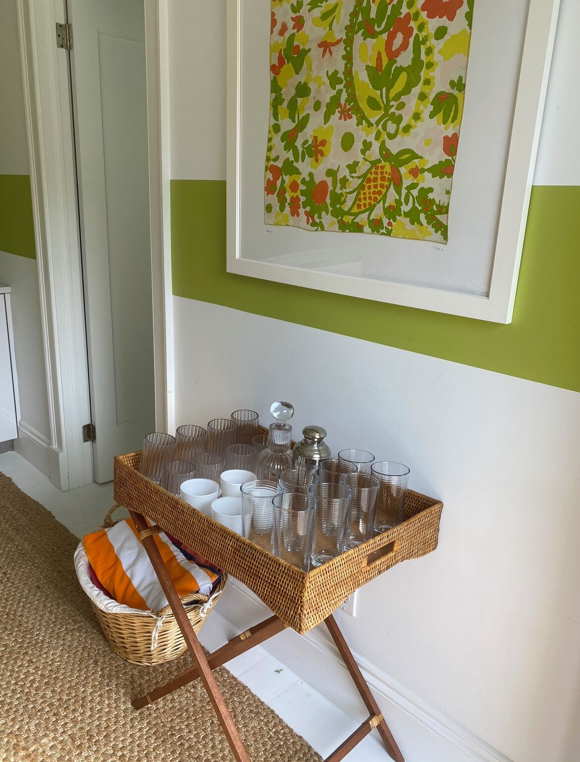 A room with a green and white wall, a framed floral artwork, and a wicker tray with glassware and a candle holder.
