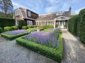 Un jardin bien entretenu, avec des haies soigneusement taillées et des plants de lavande devant une maison en briques dotée d’un toit à pignon.