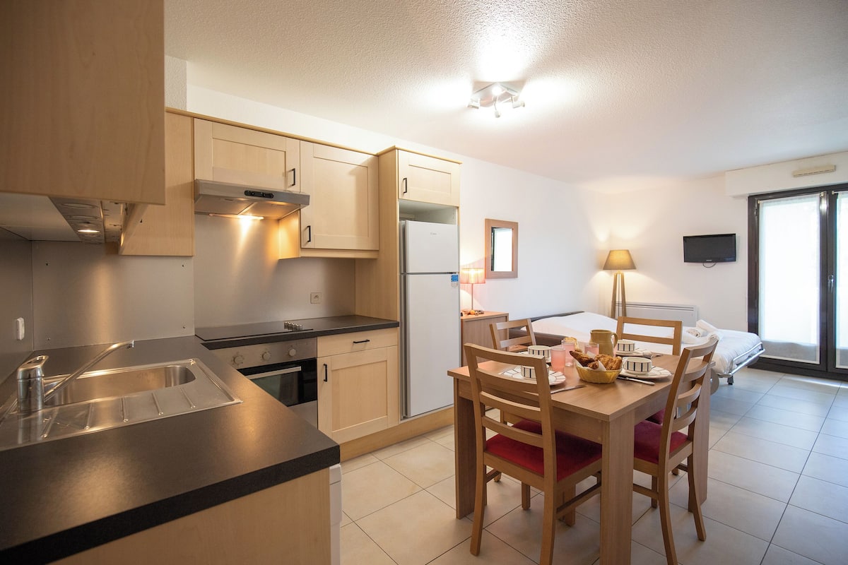 A compact kitchen with modern appliances, a dining area with wooden furniture, and a bed in the adjacent room.