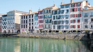Une rangée de bâtiments aux couleurs vives longe le bord de l’eau, avec un pont de pierre et des espaces de restauration en plein air.