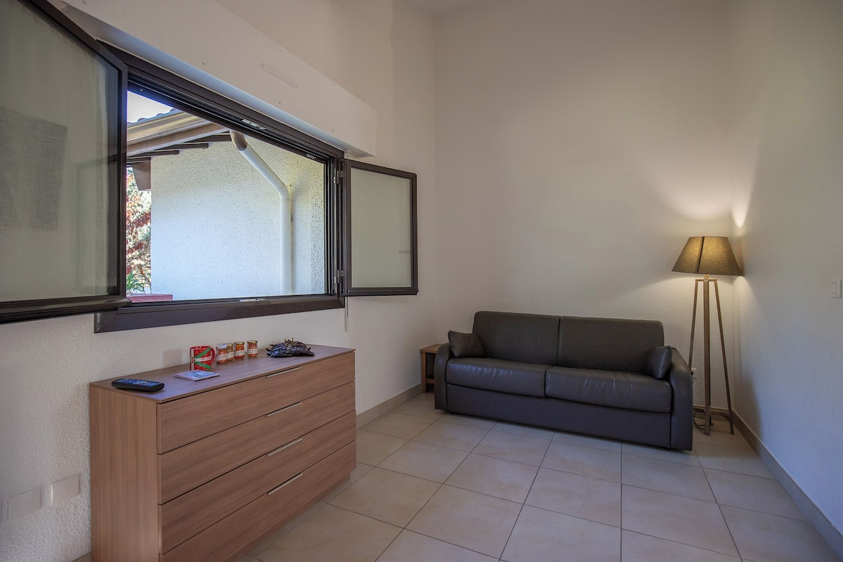 A room with a wooden dresser, a grey sofa, a floor lamp, and a window with a view of a building outside.