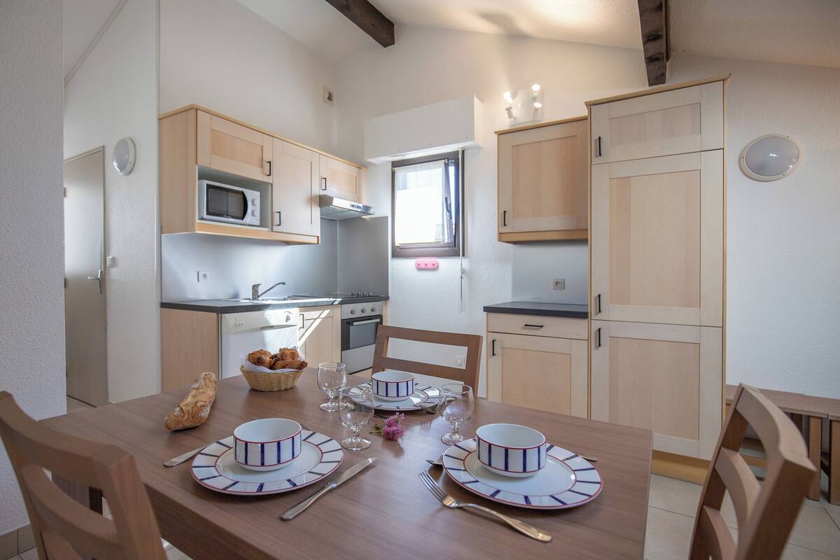 A compact kitchen with wooden cabinets, a microwave, and a dining area set for four with plates, cutlery, and glasses.