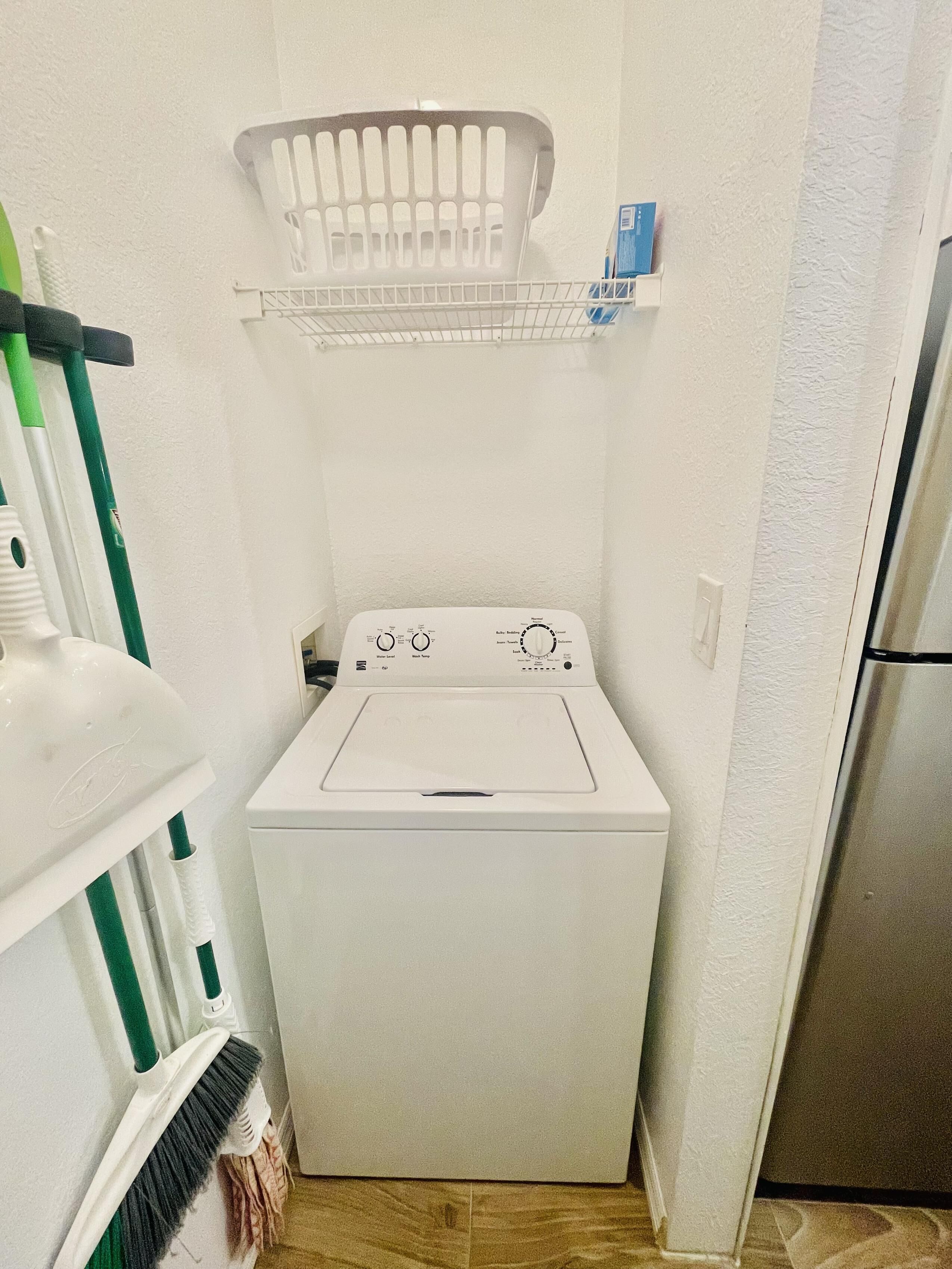 A compact laundry room with a front-loading washing machine, a drying rack, and a broom.
