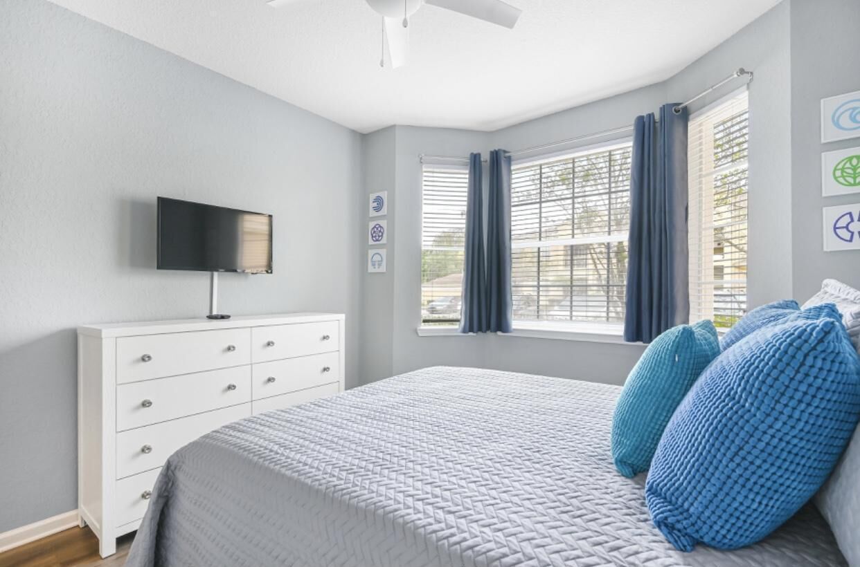 A bedroom with a bed, a TV mounted on the wall, a window with curtains, and a white dresser.