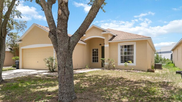 A single-story house with a beige exterior, a garage, and a large tree in the front yard. - 5/3 Pool Home Located in Sunset Ridge Community! (Davenport)