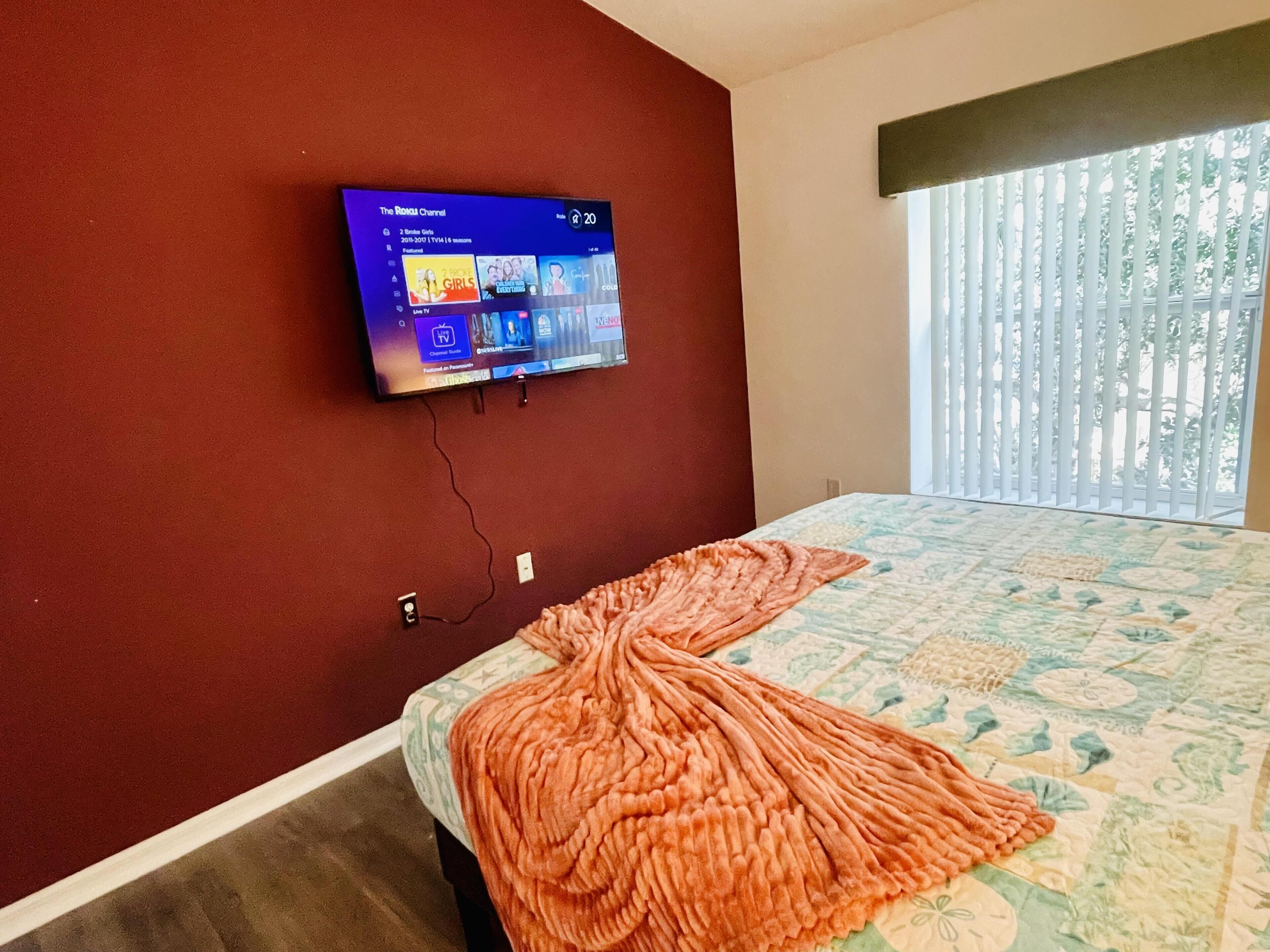 A bedroom with a bed, a TV mounted on the wall, and a window with blinds.