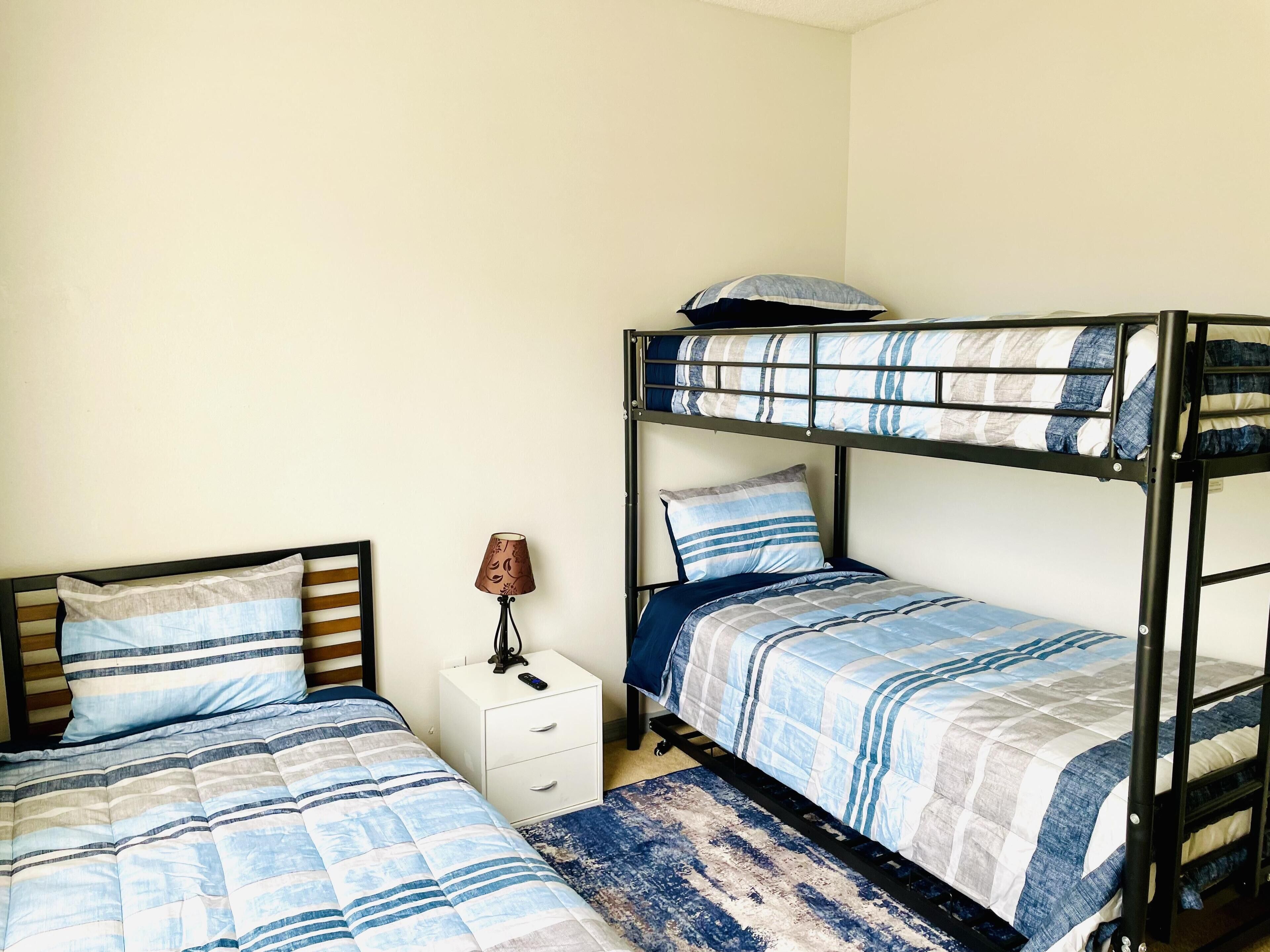 Een studentenkamer met twee eenpersoonsbedden, een stapelbed, een witte nachtkastje met lamp en een blauw-wit gestreept vloerkleed.